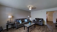 This two-story space offers a practical and comfortable layout across two levels. The living area showcases dark hardwood-style flooring that contrasts attractively with the neutral gray-toned walls, creating a clean and cohesive aesthetic. A textured popcorn ceiling adds a classic touch overhead, while a ceiling fan with an integrated light fixture provides both ambient lighting and air circulation throughout the room. The living space is generously proportioned, comfortably accommodating a full sofa and loveseat arrangement centered around a marble-style coffee table. The seating area is accented with teal and blue decorative pillows, adding pops of color against the dark upholstery. A floor lamp in the corner provides additional lighting options, enhancing the room's functionality. The open wall space and simple layout allow for flexible furniture arrangement. The overall ambiance of the living area is relaxed and welcoming, with a neutral color palette that provides a versatile backdrop. The two-floor configuration suggests additional living or sleeping space on the upper level, offering separation between common and private areas of the home.