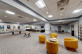 Located at 1850 Post Rd in Warwick, Rhode Island, this generously sized single-floor amenity space is designed to accommodate a wide range of social and recreational activities. The open floor plan is defined by a neutral palette of light gray walls accented with navy blue trim, patterned wall-to-wall carpeting, and a suspended tile ceiling with recessed and panel lighting throughout.

The space is thoughtfully divided into several functional zones. A game area features a pool table and foosball table, providing entertainment at the heart of the room. Adjacent to this is a casual lounge area furnished with plush dark sofas, tufted leather benches, and glass coffee tables centered around a wall-mounted flat-screen television. A second seating cluster showcases vibrant yellow upholstered swivel chairs arranged around a round side table, adding a bold pop of color to the space.

A dining and bar-height seating area with tall stools and round tables offers a more intimate setting, while a dedicated workspace includes a modern desk and ergonomic chairs, suitable for focused tasks. The space also features an accordion-style folding room divider, allowing the floor plan to be partitioned as needed. Movie posters and botanical wall art add character throughout, and a separate entry area with double doors provides direct access to the space.