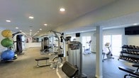 This fitness center is designed to support a comprehensive workout experience within a single-floor layout. The space features a generous open floor plan that accommodates a diverse selection of exercise equipment, including Precor cable and strength machines, seated weight stations, stationary bikes, and additional cardio equipment positioned near large windows that allow natural light to fill the room. The flooring transitions between carpet and hard surface zones, helping to delineate strength and cardio areas. Recessed lighting is evenly distributed across the smooth white ceiling, ensuring consistent illumination throughout the space. A wall of mirrors along one side visually expands the room and provides exercisers with proper form feedback during workouts. Structural columns are integrated cleanly into the layout without disrupting the flow of the space. A wall-mounted television is also present, adding convenience for users during longer training sessions. The overall ambiance is clean, functional, and well-maintained, with a neutral color palette of whites and grays lending a modern, professional feel to the environment.