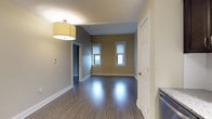 This 1-bedroom, 1-bathroom residence is thoughtfully laid out across a single floor, offering an efficient and comfortable living environment. The living area is a standout feature, showcasing rich, dark hardwood-style flooring that runs throughout and reflects the warm overhead lighting from a modern drum pendant light fixture. Two windows along the far wall allow natural light to filter in, brightening the space and creating an inviting atmosphere. Neutral-toned walls in warm greige complement the clean white trim and six-panel doors, lending a cohesive and polished aesthetic throughout.

The kitchen is positioned adjacent to the living area and is equipped with dark cabinetry, a sink, and appliances, making efficient use of the available footprint. A hallway connects the main living spaces to the bedroom and bathroom, providing a logical and private flow through the unit.

The bathroom is neatly appointed and includes a bathtub, while the bedroom offers a quiet retreat with ample space for furnishings. Throughout the unit, the consistent use of hardwood-style flooring, crisp white millwork, and neutral paint tones creates a unified and clean interior design. The single-floor layout ensures seamless accessibility and easy navigation across all rooms.
