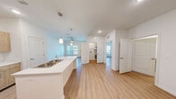 This thoughtfully designed one-bedroom, one-bathroom residence at Lalita Senior Living offers a bright, open-concept floor plan ideal for comfortable senior living. Light wood-tone flooring flows consistently throughout the space, complementing the crisp white walls and creating a clean, modern aesthetic.

The kitchen serves as a central hub of the home, anchored by a white peninsula island with a stainless steel sink and elegant pendant lighting above. Natural wood-toned cabinetry lines the walls, paired with black appliances including a refrigerator, range, and microwave. Recessed lighting adds warmth and practicality to the workspace.

The living area is bright and airy, featuring large windows that invite natural light and offer outdoor views, along with a ceiling fan for added comfort. A glass-paneled entry door further enhances the sense of openness and connectivity to the outside.

The bedroom is a well-proportioned, tranquil retreat with its own ceiling fan, large window with outdoor views, and clean white walls. The utility room provides convenient in-unit functionality.

The bathroom is designed with accessibility in mind, featuring sturdy grab bars near the shower and toilet, white subway tile in the shower area, and a warm wood-toned barn-style door — combining safety with contemporary style.