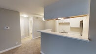 Located at 1144 Johnson Dr in Buffalo Grove, IL, this single-floor unit offers a practical and well-organized layout designed for comfortable everyday living. The open-concept kitchen features a peninsula with a blue-gray countertop, white cabinetry with under-cabinet lighting, a stainless steel refrigerator, an electric range, and a built-in sink — all flowing seamlessly into the carpeted living area. The living space is generously sized, with neutral gray walls, clean white baseboards, and a welcoming entry door that anchors the room. A dedicated laundry closet off the kitchen houses a stacked washer and dryer unit, adding exceptional in-unit convenience. The kitchen and laundry area feature wood-look flooring, while soft carpet extends throughout the living and bedroom spaces. The bathroom is finished in a clean, bright palette with tile flooring, a tub-shower combination with subway tile surround, a white vanity with an integrated sink, a large mirror, and a tall storage cabinet for added organization. The bedroom is a comfortable, carpeted room with a window fitted with horizontal blinds, allowing natural light to filter in. Throughout the unit, the consistent use of neutral tones and practical finishes creates a cohesive and move-in-ready atmosphere.