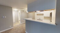 Located at 1144 Johnson Dr in Buffalo Grove, IL, this single-floor unit offers a practical and well-organized layout designed for comfortable everyday living. The open-concept kitchen features a peninsula with a blue-gray countertop, white cabinetry with under-cabinet lighting, a stainless steel refrigerator, an electric range, and a built-in sink — all flowing seamlessly into the carpeted living area. The living space is generously sized, with neutral gray walls, clean white baseboards, and a welcoming entry door that anchors the room. A dedicated laundry closet off the kitchen houses a stacked washer and dryer unit, adding exceptional in-unit convenience. The kitchen and laundry area feature wood-look flooring, while soft carpet extends throughout the living and bedroom spaces. The bathroom is finished in a clean, bright palette with tile flooring, a tub-shower combination with subway tile surround, a white vanity with an integrated sink, a large mirror, and a tall storage cabinet for added organization. The bedroom is a comfortable, carpeted room with a window fitted with horizontal blinds, allowing natural light to filter in. Throughout the unit, the consistent use of neutral tones and practical finishes creates a cohesive and move-in-ready atmosphere.