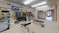 This fitness center offers a thorough and thoughtfully organized workout environment spread across a single open floor. The space is defined by a striking architectural feature: tall, stacked natural wood columns that rise from floor to ceiling, lending a warm, modern aesthetic that contrasts with the neutral tones of the carpet flooring and light gray walls. Bright overhead fluorescent lighting ensures the entire space is well-illuminated throughout.

The cardio zone is well-stocked with multiple treadmills, elliptical machines, a rowing machine, and a stationary bike, each positioned for ample spacing between users. Several wall-mounted flat-screen televisions are placed throughout the area, allowing members to stay entertained during workouts.

The strength training area features a full rack of dumbbells, adjustable weight benches, a cable/functional trainer machine, and a vertical knee raise station, providing options for both free weight and machine-based training. A large wall mirror in the free weights section enhances the sense of space and allows for form monitoring.

A glass-enclosed private studio room is accessible through sleek glass sliding doors. This separate area is visible from the main floor and appears suited for personal training, group classes, or focused individual workouts. The overall ambiance of the fitness center is clean, modern, and functional, catering to a wide range of fitness needs.