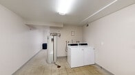 This single-floor shared laundry facility is a practical, well-organized utility space divided into two functional rooms. The main laundry room features light-toned walls, speckled vinyl tile flooring, and flush-mounted ceiling lighting that provides even illumination throughout. The room houses multiple top-load washing machines and dryers positioned along the wall, accompanied by plumbing hookups and a large standing water heater. A laundry cart is available for residents' convenience, and posted notices on the wall provide usage guidelines. Ventilation is ensured through ceiling-mounted HVAC vents. The second room serves as an anteroom or multipurpose area, featuring the same speckled flooring, neutral walls, and overhead lighting. It includes a folding table and is equipped with a wall-mounted exhaust fan. A standard interior door connects the two rooms, while a prominent blue exterior door provides direct access to an outdoor covered walkway, making entry and exit highly convenient. The overall design is clean, utilitarian, and efficiently laid out to support the day-to-day laundry needs of a multi-unit residential building.