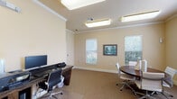 This single-floor business center is thoughtfully designed to accommodate a range of professional needs. The space is finished with warm beige walls, crown molding, and neutral carpet throughout, creating a polished and focused atmosphere. Recessed fluorescent ceiling fixtures provide bright, even illumination across the entire area.

The workstation zone features a long dark granite-topped desk accommodating multiple computer terminals, complete with ergonomic rolling chairs and a shared printer, making it well-suited for productivity tasks. A dark wood bookcase stocked with reference materials adds a practical and classic touch to this area.

A glass-paneled interior French door leads to a dedicated meeting area, offering a degree of privacy while maintaining visual openness. This meeting space is furnished with a round dark wood conference table surrounded by modern white chairs, ideal for small group discussions or collaborative sessions. Multiple windows dressed with horizontal blinds line the walls, allowing natural light to filter in and providing views of the surrounding greenery. A framed landscape painting serves as a tasteful focal point on one wall.

The overall design balances functionality with a comfortable, professional ambiance, making this business center a versatile and inviting workspace.