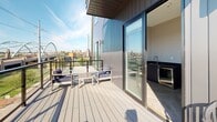 Located in Providence, Rhode Island, this contemporary single-floor space combines modern design with practical functionality and impressive outdoor access. The interior is characterized by clean lines, neutral tones, and an open layout that connects the living, sleeping, and dining areas seamlessly. The living area features a large sectional sofa in soft blue-gray tones, accented with decorative pillows, and is equipped with a wall-mounted climate control unit for year-round comfort. The dining area is furnished with a round dark-wood table and matching chairs, set against light herringbone-pattern hardwood flooring. A compact kitchenette with a dark countertop and sink adds practical utility to the space. Glass-paneled doors with dark frames open to the building's hallway, offering a polished, modern aesthetic. The standout feature of this space is its generous outdoor deck, accessible via large sliding glass doors. The deck is outfitted with outdoor seating and an exterior sink, making it ideal for entertaining. Glass railings ensure unobstructed panoramic views of the Providence waterfront, surrounding greenery, and the city skyline beyond. The combination of contemporary interiors, thoughtful amenities, and exceptional outdoor space makes this a distinctive urban retreat in the heart of Providence.