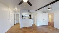 This single-story unit offers a thoughtfully designed open-concept layout that maximizes the sense of space throughout. The main living area features vaulted ceilings accented by a dark exposed beam, adding architectural character to the bright, airy interior. A ceiling fan with a sleek dark finish is centrally positioned, providing both style and function. Warm-toned hardwood flooring runs throughout, creating a cohesive and inviting feel.

The kitchen is semi-open, defined by a partial wall that serves as a breakfast bar with a dark countertop. White cabinetry with glass-front upper doors provides ample storage, while stainless steel appliances—including a refrigerator and microwave—offer modern functionality. The kitchen's connection to the living area promotes an easy flow ideal for everyday living.

Multiple doorways visible from the main space lead to additional rooms, including what appears to be a well-sized bedroom and a dedicated storage or utility area with built-in shelving. Crisp white walls throughout reflect natural and ambient light, enhancing the open, clean aesthetic of the unit.