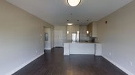 This single-floor unit offers a clean, contemporary design centered around an open-concept living and kitchen area. The kitchen is a standout feature, boasting light-toned cabinetry, a white kitchen island with an integrated sink, and stainless steel appliances including a refrigerator and range. Three stylish pendant lights hang above the island, complementing the flush-mount ceiling fixture that illuminates the main living space. Soft, neutral-toned walls create a bright and airy atmosphere throughout, while rich, dark hardwood flooring grounds the space with warmth and contrast. The open layout allows for a seamless flow between the kitchen and living areas, providing a versatile and functional floor plan. A hallway visible from the main area leads to additional rooms, suggesting a well-organized and private layout. An electrical panel is conveniently recessed into the wall near the entryway corridor. The overall ambiance is modern and minimalist, making it an ideal canvas for a variety of interior design styles.