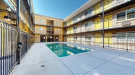 Located at 291 W Belleview Ave in Englewood, Colorado, this outdoor courtyard pool area is a standout communal amenity set within a multi-story residential building. The space is defined by a centrally positioned rectangular swimming pool with clear, blue-green water, surrounded by a generous expanse of smooth concrete decking that provides ample room for lounging and recreation.

The pool is enclosed by black metal safety fencing, creating a secure and well-defined boundary. The surrounding building features a classic brick facade across three stories, accented by vibrant yellow doors and railings that lend a bold, modern aesthetic to the otherwise traditional exterior. Open-air walkways and balconies line each level, offering residents direct sightlines into the courtyard below.

Access between floors is provided via metal staircases visible at the corners of the building, and ceiling-mounted lighting fixtures are positioned along the covered walkways to illuminate the space after dark. The courtyard orientation allows for abundant natural light throughout the day, with open sky above providing an airy, open-air atmosphere. The combination of the bright yellow accents against the textured brick exterior creates a visually energetic environment, making this pool courtyard a vibrant and functional centerpiece of the property.