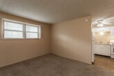 This single-floor unit offers a straightforward, functional layout designed for comfortable everyday living. The living area is finished with wall-to-wall carpet and neutral-toned walls, with a window fitted with blinds that allows natural light to fill the space. A textured ceiling adds subtle visual character throughout the main areas. The layout flows directly into the kitchen, which features light blue cabinetry, a double-basin sink, an electric range, and a full-size refrigerator. Tile flooring and a ceiling fan with an integrated light fixture give the kitchen a bright, practical feel. Adjacent to the living space, a bedroom area includes mirrored sliding closet doors that reflect light and contribute to a sense of openness, along with a secondary door providing additional storage or access. The full bathroom is finished with white wall tiles accented by dark red tile details, a soaking tub, a toilet, and a single-sink vanity with cabinet storage. A mosaic tile floor and a window with blinds complete the bathroom's clean, utilitarian aesthetic. Throughout the unit, ceiling fans, built-in ventilation, and multiple electrical outlets provide convenience and comfort.