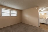 This single-floor unit offers a straightforward, functional layout designed for comfortable everyday living. The living area is finished with wall-to-wall carpet and neutral-toned walls, with a window fitted with blinds that allows natural light to fill the space. A textured ceiling adds subtle visual character throughout the main areas. The layout flows directly into the kitchen, which features light blue cabinetry, a double-basin sink, an electric range, and a full-size refrigerator. Tile flooring and a ceiling fan with an integrated light fixture give the kitchen a bright, practical feel. Adjacent to the living space, a bedroom area includes mirrored sliding closet doors that reflect light and contribute to a sense of openness, along with a secondary door providing additional storage or access. The full bathroom is finished with white wall tiles accented by dark red tile details, a soaking tub, a toilet, and a single-sink vanity with cabinet storage. A mosaic tile floor and a window with blinds complete the bathroom's clean, utilitarian aesthetic. Throughout the unit, ceiling fans, built-in ventilation, and multiple electrical outlets provide convenience and comfort.