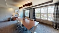 This well-appointed conference room offers a polished, professional environment designed to accommodate productive meetings and gatherings. At the center of the space sits a substantial rich wood boardroom table surrounded by approximately ten white upholstered executive chairs with chrome bases, providing a clean, contemporary aesthetic. The room is anchored by a striking cluster of sculptural drum pendant lights in warm gold and bronze tones, suspended from a tray ceiling that adds architectural depth and dimension overhead.

One wall features a large wall-mounted flat-screen television, ideal for presentations, while the opposite end of the room is adorned with two bold abstract art pieces in muted yellows, whites, and grays, lending a refined artistic touch. A wood credenza along one wall provides additional surface space and storage. Patterned gray carpeting runs throughout, grounding the room with texture and warmth.

Large windows dressed with dark plaid drape panels line one side of the room, allowing generous natural light to fill the space while offering views of a landscaped exterior courtyard and surrounding buildings. The overall ambiance is contemporary and sophisticated, balancing functional meeting space with thoughtful design details.