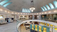 Located at 114 SE Douglas St in Lee's Summit, Missouri, this thoughtfully renovated historic space masterfully blends classical architectural grandeur with contemporary functionality. The centerpiece of the interior is a soaring domed rotunda with a coffered, vaulted ceiling finished in soft gray tones, crowned by a elegant multi-globe chandelier suspended from an ornate cross-beam. A continuous clerestory of arched windows encircles the upper dome, flooding the space with diffused natural light and creating an airy, uplifting atmosphere.

A curved glass-railed mezzanine wraps around the rotunda's upper level, offering open-plan workspace with long work tables and flexible seating arrangements. The lower level features a welcoming reception area with a dark wood desk, neutral-toned lounge chairs, and warm hardwood and textured tile flooring. Illuminated stained glass panels in amber and teal tones line accent walls, adding a warm, vibrant character to the interior.

A glass-enclosed conference room offers a private meeting space with a round table, ergonomic seating, arched windows, and hardwood floors. Additional lounge and collaborative seating areas are thoughtfully distributed throughout the space. The overall ambiance is professional yet inviting, honoring the building's historic character while serving modern needs.