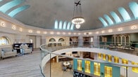 Located at 114 SE Douglas St in Lee's Summit, Missouri, this thoughtfully renovated historic space masterfully blends classical architectural grandeur with contemporary functionality. The centerpiece of the interior is a soaring domed rotunda with a coffered, vaulted ceiling finished in soft gray tones, crowned by a elegant multi-globe chandelier suspended from an ornate cross-beam. A continuous clerestory of arched windows encircles the upper dome, flooding the space with diffused natural light and creating an airy, uplifting atmosphere.

A curved glass-railed mezzanine wraps around the rotunda's upper level, offering open-plan workspace with long work tables and flexible seating arrangements. The lower level features a welcoming reception area with a dark wood desk, neutral-toned lounge chairs, and warm hardwood and textured tile flooring. Illuminated stained glass panels in amber and teal tones line accent walls, adding a warm, vibrant character to the interior.

A glass-enclosed conference room offers a private meeting space with a round table, ergonomic seating, arched windows, and hardwood floors. Additional lounge and collaborative seating areas are thoughtfully distributed throughout the space. The overall ambiance is professional yet inviting, honoring the building's historic character while serving modern needs.