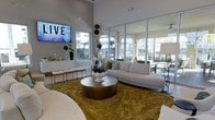 This single-floor space showcases a sophisticated, open-plan lounge area defined by its impressive ceiling height and expansive floor-to-ceiling glass windows and doors. The generous glazing floods the interior with natural light while offering views of an outdoor terrace and surrounding landscaping, creating a seamless connection between indoor and outdoor environments.

The lounge is furnished with a curved, neutral-toned sectional sofa accented with decorative throw pillows, anchored by a substantial metallic drum coffee table atop a textured gold area rug. A wall-mounted flat-screen television serves as a focal point on one side of the room, accompanied by a sleek white credenza styled with decorative accessories and greenery. A striking tall floral and branch arrangement adds vertical interest and organic texture to the space.

The overall aesthetic is clean and contemporary, combining white walls and gray-toned architectural framing with warm metallic and natural accents. Seating is versatile and abundant, with additional bench seating along the perimeter. The space conveys an upscale, welcoming ambiance suited for lounging or socializing.