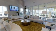 This single-floor space showcases a sophisticated, open-plan lounge area defined by its impressive ceiling height and expansive floor-to-ceiling glass windows and doors. The generous glazing floods the interior with natural light while offering views of an outdoor terrace and surrounding landscaping, creating a seamless connection between indoor and outdoor environments.

The lounge is furnished with a curved, neutral-toned sectional sofa accented with decorative throw pillows, anchored by a substantial metallic drum coffee table atop a textured gold area rug. A wall-mounted flat-screen television serves as a focal point on one side of the room, accompanied by a sleek white credenza styled with decorative accessories and greenery. A striking tall floral and branch arrangement adds vertical interest and organic texture to the space.

The overall aesthetic is clean and contemporary, combining white walls and gray-toned architectural framing with warm metallic and natural accents. Seating is versatile and abundant, with additional bench seating along the perimeter. The space conveys an upscale, welcoming ambiance suited for lounging or socializing.