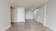 This single-floor unit offers a clean, functional layout designed for practical living. The main living area is an open, well-proportioned space finished with grey wood-look vinyl plank flooring and neutral grey walls, creating a bright and cohesive atmosphere. A large window allows natural light to filter in, and a baseboard heating system along with a wall-mounted air conditioning unit ensures year-round climate control.

The kitchen is efficiently arranged and equipped with black appliances, including an electric range with overhead microwave, a full-size refrigerator, and a dishwasher. Warm-toned wooden cabinetry pairs with light stone-finish countertops, offering ample storage and prep space.

The bedroom features soft carpeting in a neutral tone, providing a comfortable contrast to the hard flooring found throughout the rest of the unit. Its simple, understated design offers a versatile space adaptable to various furniture arrangements.

The bathroom is well-appointed with a full soaking tub and shower combination framed in a clean white surround, complemented by wood-finish cabinetry beneath a light countertop vanity. Matching vinyl plank flooring ties the bathroom seamlessly to the rest of the unit's design. Overall, the space presents a consistent, move-in-ready aesthetic throughout.