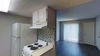 This single-floor space offers an efficient, open layout connecting the kitchen and living area. The kitchen is equipped with a white coil-burner electric range, light-toned laminate countertops, and white upper cabinetry with a built-in range hood. A distinctive mosaic tile backsplash in muted gray and brown tones adds a decorative focal point above the stove. The textured popcorn ceiling is consistent throughout the space, and the walls feature a subtle textured finish in a neutral gray tone. The kitchen transitions naturally into the living area, which features dark hardwood-style flooring that contrasts with the lighter walls and ceiling. A large window dressed with vertical blinds allows natural light to fill the living space, contributing to an open and airy atmosphere. The overall design blends practical functionality with modest contemporary touches, making the space well-suited for everyday living. The open sightline between the kitchen and living room enhances the sense of spaciousness throughout this single-level unit.