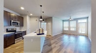 This single-floor unit offers a well-designed, open-concept layout that seamlessly connects the kitchen and living area. The kitchen is a standout feature, anchored by a large center island with a white countertop and an undermount sink, illuminated by a pair of sleek black pendant lights. Dark espresso cabinetry extends to the ceiling, providing ample storage, while a subway tile backsplash adds a clean, contemporary touch. Stainless steel appliances — including a refrigerator with ice and water dispenser, electric range, and built-in microwave — complete the space.

Light wood-style plank flooring runs throughout the main living areas, contributing to a bright, cohesive aesthetic. The open living area benefits from natural light streaming through multiple windows fitted with horizontal blinds, and a ceiling fan provides additional comfort. Bedrooms feature plush carpeting and corner window configurations that invite generous natural light.

The bathroom is finished with white subway tile surrounding a full tub-shower combination, paired with a dark vanity and light countertop. A dedicated laundry closet houses a full-size, top-load washer and dryer with built-in shelving above. Neutral gray walls and clean white trim unify the space throughout.