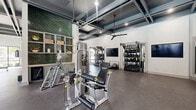 This professionally designed fitness center offers a generous, open floor plan thoughtfully organized into distinct training zones. The space is defined by its striking coffered ceilings with dark-painted beams, recessed lighting, and large ceiling fans that enhance airflow and comfort throughout.

The cardio zone runs along a wall of floor-to-ceiling windows, flooding the area with natural light and offering outdoor views. A row of treadmills and elliptical machines lines this bright corridor, creating an energizing environment for aerobic workouts.

The strength training area is comprehensively outfitted with cable machines, multi-functional racks, adjustable benches, and a full dumbbell rack with an accompanying flat-screen TV. A decorative green subway tile accent wall adds a bold visual element behind built-in shelving, lending a boutique gym aesthetic to the space.

A dedicated functional training zone features TRX suspension systems, battle ropes, slam balls, and open floor space for dynamic movement. Sliding barn-style doors connect seamlessly to an adjacent area, allowing flexible use of the layout.

Throughout the facility, durable rubber flooring provides safety and comfort underfoot, while white walls and abundant natural light maintain a clean, modern atmosphere. The overall design balances high-end aesthetics with practical, performance-focused functionality.