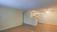 This single-floor residence offers a practical and well-organized layout designed for comfortable everyday living. The open-concept main area seamlessly connects the living space to the kitchen, creating a sense of openness and ease of movement throughout. Warm hardwood-style flooring runs continuously across the main living area, adding a touch of warmth and cohesion to the space.

The kitchen is positioned at one end of the main room, defined by a half-wall peninsula that provides both visual separation and functional counter space. White cabinetry lines the kitchen walls, offering ample storage, while white appliances—including a full-size refrigerator—contribute to a clean, uniform aesthetic. A sink and additional counter space complete the kitchen's efficient setup.

The living area is generously sized and bathed in soft, ambient light, with neutral yellow-toned walls that create a welcoming atmosphere. A textured ceiling adds a subtle architectural detail throughout. A doorway off the main living area leads to additional rooms, suggesting a thoughtful floor plan that balances open communal spaces with more private areas. Overall, the space presents a straightforward, move-in-ready environment with a light and airy feel.