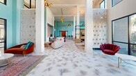 This expansive single-floor amenity space makes a striking first impression with its soaring double-height ceilings, bold teal-painted walls, and an abundance of natural light streaming through floor-to-ceiling black-framed windows. The patterned tile flooring flows throughout, unifying the various distinct zones within the open layout.

Upon entry, a welcoming foyer features sculptural red-orange lounge chairs set against a textured 3D white accent wall, creating an eye-catching vignette alongside built-in bench seating. Large glass doors offer direct views of the landscaped outdoor surroundings, including palm trees and greenery.

The main lounge area is anchored by generous sectional sofas in neutral tones accented with vibrant throw pillows, centered around a gold cocktail table beneath a statement wall-mounted television. Decorative metallic screen partitions frame the space elegantly, while pendant chandeliers cast warm ambient light throughout.

A co-working bar counter outfitted with a mix of colorful swivel stools and a mounted display screen provides a functional workspace. Moving deeper into the space, a teal-walled social lounge and game room features high communal tables, bar-height seating, pendant globe lighting, and a pool table. Vibrant large-scale artwork punctuates the walls throughout. A dedicated indoor golf simulator room with a turf mat and projection screen rounds out this well-appointed amenity suite.