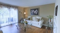 This single-floor space offers a welcoming and well-appointed interior defined by its warm neutral color palette and classic design sensibility. The living area serves as the heart of the home, showcasing rich hardwood flooring that adds warmth and character throughout. Large windows dressed with elegant vertical blinds and decorative curtains flood the room with natural light, creating an airy and open atmosphere.

The living room is thoughtfully arranged with a plush white slipcovered sofa accented by earth-toned throw pillows, centered around a distinctive glass-top coffee table with an ornate metal base. A framed botanical artwork on the warm beige wall adds a refined decorative touch, complemented by a classic floor lamp that provides soft ambient lighting. A decorative lantern with candle accents and a fresh floral arrangement on the coffee table enhance the inviting ambiance.

White baseboards and clean wall finishes contribute to a tidy, well-maintained aesthetic throughout. The natural light streaming in from what appears to be a sliding glass door or large window hints at outdoor access, adding to the sense of space and connectivity to the exterior. Overall, this space presents a cozy, classically styled interior with practical and comfortable living in mind.