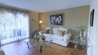 This single-floor space offers a welcoming and well-appointed interior defined by its warm neutral color palette and classic design sensibility. The living area serves as the heart of the home, showcasing rich hardwood flooring that adds warmth and character throughout. Large windows dressed with elegant vertical blinds and decorative curtains flood the room with natural light, creating an airy and open atmosphere.

The living room is thoughtfully arranged with a plush white slipcovered sofa accented by earth-toned throw pillows, centered around a distinctive glass-top coffee table with an ornate metal base. A framed botanical artwork on the warm beige wall adds a refined decorative touch, complemented by a classic floor lamp that provides soft ambient lighting. A decorative lantern with candle accents and a fresh floral arrangement on the coffee table enhance the inviting ambiance.

White baseboards and clean wall finishes contribute to a tidy, well-maintained aesthetic throughout. The natural light streaming in from what appears to be a sliding glass door or large window hints at outdoor access, adding to the sense of space and connectivity to the exterior. Overall, this space presents a cozy, classically styled interior with practical and comfortable living in mind.