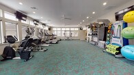 This single-level fitness center offers a comprehensive workout environment designed to accommodate a wide range of exercise needs. The space is generously proportioned, with an open floor plan that allows for easy movement between equipment zones. Underfoot, a distinctive teal and brown patterned carpet tile covers the entire floor, providing both cushioning and visual appeal throughout the room.

The cardio section lines one wall and features a row of treadmills, elliptical trainers, and stair climbers, each positioned in front of large windows that allow natural light to fill the space and offer exterior views. Wall-mounted flat-screen televisions are positioned at intervals above the cardio equipment, enabling users to watch content during their workouts.

On the opposite side of the room, a well-organized functional training station houses TRX suspension trainers, resistance bands, medicine balls, and stability balls in multiple sizes. A dedicated free weights area includes a dumbbell rack and adjustable bench, while a multi-station cable machine supports a variety of strength exercises.

Ceiling fans and recessed lighting are distributed evenly throughout, ensuring consistent airflow and illumination. The neutral wall tones and white ceiling contribute to a clean, bright atmosphere. A secondary door provides additional access to the space, enhancing circulation and convenience.
