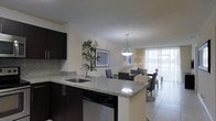 This single-floor unit showcases a thoughtfully designed open-concept layout that seamlessly connects the kitchen, dining, and living areas into one cohesive living space. The kitchen is outfitted with dark espresso cabinetry that provides a striking contrast against the light speckled granite countertops, which extend across a peninsula breakfast bar — ideal for casual dining or meal prep. A stainless steel undermount sink and dishwasher add a contemporary, functional touch. A stylish pendant light hangs above the peninsula, anchoring the kitchen visually. The dining area sits just beyond the kitchen and comfortably accommodates a full dining set with upholstered chairs. The living room benefits from an abundance of natural light streaming through floor-to-ceiling windows dressed with vertical blinds, creating a bright and airy atmosphere. Light-toned tile flooring runs throughout, enhancing the sense of openness. Neutral wall tones provide a versatile backdrop, while decorative elements such as framed artwork and indoor plants add warmth and character. Overall, the space offers a clean, modern aesthetic well-suited for everyday living.