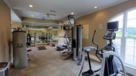 This single-floor fitness center is designed to support a well-rounded workout routine. The space is outfitted with a variety of exercise equipment, including treadmills, an elliptical trainer, a multi-station strength training unit, and additional free-standing machines, providing options for both cardiovascular and resistance training. The carpeted flooring adds a degree of comfort underfoot, while a large mirrored wall along one side visually expands the room and allows users to monitor their form during exercise. Recessed lighting is installed throughout the ceiling, ensuring consistent, even illumination across the workout area. Ceiling fans provide additional airflow, contributing to a more comfortable environment during exercise. Crown molding along the upper walls adds a subtle architectural detail, lending the space a polished finish. The warm, neutral-toned walls create a focused yet approachable atmosphere. A posted fitness center rules notice is visible, indicating this is a shared community or facility amenity space. Overall, the layout is functional and efficiently organized to maximize usable floor space.