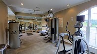 This single-floor fitness center is designed to support a well-rounded workout routine. The space is outfitted with a variety of exercise equipment, including treadmills, an elliptical trainer, a multi-station strength training unit, and additional free-standing machines, providing options for both cardiovascular and resistance training. The carpeted flooring adds a degree of comfort underfoot, while a large mirrored wall along one side visually expands the room and allows users to monitor their form during exercise. Recessed lighting is installed throughout the ceiling, ensuring consistent, even illumination across the workout area. Ceiling fans provide additional airflow, contributing to a more comfortable environment during exercise. Crown molding along the upper walls adds a subtle architectural detail, lending the space a polished finish. The warm, neutral-toned walls create a focused yet approachable atmosphere. A posted fitness center rules notice is visible, indicating this is a shared community or facility amenity space. Overall, the layout is functional and efficiently organized to maximize usable floor space.