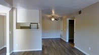This single-floor space features an open-concept layout that seamlessly connects the kitchen and living areas. Dark hardwood-style flooring runs throughout, providing a warm, cohesive foundation that contrasts nicely with the neutral, light-toned walls and white trim.

The kitchen is partially separated from the living area by a half-wall pass-through, which includes a countertop surface and overhead cabinetry. A microwave is integrated into the upper cabinetry, offering convenient functionality. This design allows for visual connectivity between the kitchen and the main living space while maintaining a subtle sense of division.

The living area is generously proportioned, with a ceiling fan centrally mounted to provide both lighting and air circulation. The textured ceiling adds a subtle architectural detail to the otherwise clean and minimalist aesthetic.

A doorway visible from the main living space leads to what appears to be a sizable walk-in closet, offering substantial storage capacity. Clean white baseboards and door trim provide a crisp finish throughout. Overall, the space presents a practical, well-maintained interior with a straightforward, functional design.