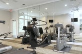This fitness center is thoughtfully designed to support a well-rounded workout experience. The space is organized into distinct functional zones, beginning with a dedicated cardio area that houses multiple Cybex treadmills, each positioned beneath wall-mounted flat-screen televisions for an engaging workout experience. Adjacent to the cardio section, a multi-station strength training unit provides a comprehensive range of resistance exercises. A rowing machine and stationary bike add further variety to the equipment lineup. Floor-to-ceiling mirrors line one wall, visually expanding the space while allowing users to monitor their form during exercise. The open exercise area features patterned modular carpet tiles over hardwood flooring, creating a practical and visually appealing surface ideal for stretching, yoga, or use of the stability ball on hand. Recessed lighting and a ceiling fan provide a comfortable and well-lit environment. Warm neutral tones on the walls are complemented by crown molding and clean white trim, giving the space a polished, upscale feel. Large French doors with transom windows along two walls flood the interior with natural light and offer views of lush greenery and an outdoor pool area, creating an inviting and energizing atmosphere throughout.