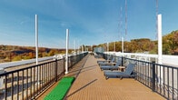 This expansive rooftop terrace offers an elevated outdoor retreat designed for relaxation and socializing. The deck is finished with warm-toned wood-plank flooring that runs the length of the rooftop, creating a cohesive and inviting surface throughout. Sturdy black metal railings line the perimeter, providing both safety and unobstructed sightlines to the surrounding landscape.

The terrace is thoughtfully furnished with multiple padded chaise lounges paired with small side tables, ideal for sunbathing or unwinding in the open air. Separate dining areas featuring wooden tables and chairs are positioned to take advantage of the sweeping views, making them well-suited for outdoor meals or casual gatherings. A compact putting green adds a recreational element to the space.

Tall white poles are installed at regular intervals along the deck, likely supporting shade structures or lighting fixtures. The rooftop enjoys abundant natural sunlight and commanding views of a lush tree canopy displaying vibrant autumn foliage in rich shades of orange, red, and green. The open sky above enhances the airy, expansive feel of the space. A utility structure on the rooftop provides additional architectural definition. Overall, this rooftop terrace combines practical amenities with a scenic, open-air environment.