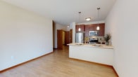 This single-floor unit is thoughtfully designed with an open, flowing layout that connects the kitchen and living areas seamlessly. Light hardwood flooring runs throughout the main living space, creating a warm and cohesive aesthetic complemented by neutral wall tones and clean white baseboards.

The kitchen is a standout feature, equipped with rich dark-wood cabinetry, a full suite of stainless steel appliances including a refrigerator, electric range, and built-in microwave, and a light-toned countertop that extends into a peninsula. Pendant lighting hangs above the peninsula, adding both task illumination and a modern design touch. The peninsula provides additional prep and dining space, making the kitchen both functional and social.

The living area is open and spacious, with a large window that invites natural light into the room. A wall-mounted climate control unit is also present, providing convenient temperature management.

The bathroom is well-appointed with a granite-topped vanity featuring dark wood cabinetry, a full-size soaking tub with shower, and brushed nickel hardware throughout. Neutral tile flooring and ample mirror space complete the practical yet refined feel of the room.

Overall, the unit offers a clean, modern interior with consistent material choices that lend the space a cohesive and comfortable character.