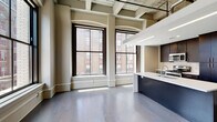 This single-floor unit showcases a compelling blend of historic architectural details and modern design elements. Throughout the open living and kitchen area, dramatic ceiling heights are accentuated by exposed structural beams and industrial-style ductwork, lending the space a distinctive loft character. Large, arched windows with dark frames line multiple walls, flooding the interior with abundant natural light and offering views of the surrounding urban streetscape. Wide-plank hardwood flooring in a cool gray tone anchors the open-plan living area.

The kitchen is a standout feature, centered around a substantial island with a crisp white countertop, an undermount sink, and a bold navy base. Dark espresso cabinetry lines the walls and reaches toward the ceiling, paired with stainless steel appliances including a refrigerator, range, and microwave.

The bedrooms transition to plush carpeting and maintain the same generously arched windows, creating bright, comfortable retreats. A dedicated bathroom features dark tile flooring, a dark vanity with a white countertop, a combined tub and shower, and a sliding frosted-glass door, offering a clean, contemporary aesthetic throughout.
