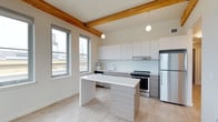This single-floor space showcases a thoughtfully designed open-concept layout that seamlessly blends industrial character with contemporary finishes. Exposed natural wood beams run across the ceiling, adding warmth and architectural interest that contrasts beautifully with the otherwise clean, modern aesthetic. Pendant globe lights hang from the beams, providing ambient illumination that complements the generous natural light streaming through the tall, multi-pane windows along the exterior wall.

The kitchen is the focal point of the space, featuring sleek flat-front upper cabinetry in crisp white paired with lower cabinets in a warm wood-tone laminate finish. Stainless steel appliances, including a refrigerator and range with an integrated hood vent, provide a polished, functional cooking environment. A white backsplash and countertops maintain the clean, minimalist palette throughout.

A central kitchen island with a white surface and wood-tone base offers additional workspace and casual dining potential. Light-toned hardwood-style flooring runs throughout, enhancing the sense of openness and warmth. The large windows frame views of the surrounding urban landscape, grounding the space within its city context.