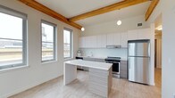 This single-floor space showcases a thoughtfully designed open-concept layout that seamlessly blends industrial character with contemporary finishes. Exposed natural wood beams run across the ceiling, adding warmth and architectural interest that contrasts beautifully with the otherwise clean, modern aesthetic. Pendant globe lights hang from the beams, providing ambient illumination that complements the generous natural light streaming through the tall, multi-pane windows along the exterior wall.

The kitchen is the focal point of the space, featuring sleek flat-front upper cabinetry in crisp white paired with lower cabinets in a warm wood-tone laminate finish. Stainless steel appliances, including a refrigerator and range with an integrated hood vent, provide a polished, functional cooking environment. A white backsplash and countertops maintain the clean, minimalist palette throughout.

A central kitchen island with a white surface and wood-tone base offers additional workspace and casual dining potential. Light-toned hardwood-style flooring runs throughout, enhancing the sense of openness and warmth. The large windows frame views of the surrounding urban landscape, grounding the space within its city context.