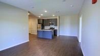 This single-floor space showcases an open-concept layout that seamlessly connects the kitchen and living areas. The kitchen is a standout feature, equipped with stainless steel appliances including a refrigerator, range, and over-the-range microwave. Dark gray cabinetry lines the walls, providing ample storage, while a central island with a contrasting blue-gray finish serves as both a workspace and a casual gathering point. Light-toned countertops complement the cabinetry, and recessed lighting illuminates the space evenly throughout.

The flooring consists of dark hardwood-style planks that extend continuously across the open area, lending warmth and cohesion to the overall design. The neutral, light gray walls create a clean backdrop that allows the kitchen's darker tones to stand out. A door visible along the far wall suggests access to additional rooms or storage. The open layout between the kitchen and the adjacent living area promotes a sense of spaciousness, making the space feel connected and functional. Overall, the design balances modern finishes with a practical, livable aesthetic.