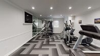 This single-floor fitness space in Greenville, South Carolina, is thoughtfully designed to deliver a comprehensive workout environment with a polished, modern aesthetic. The room is finished with crisp white walls accented by a classic chair-rail molding detail that runs the perimeter, lending the space a refined, structured appearance. Crown molding at the ceiling adds an additional architectural touch, elevating the overall design beyond a typical utility room.

Recessed lighting is evenly distributed across the smooth white ceiling, providing bright, consistent illumination throughout the space. A wall-mounted flat-screen television is positioned for easy viewing during workouts, adding a functional entertainment element. Full-length mirrors along one wall visually expand the room while serving practical fitness purposes.

The flooring consists of geometric patterned carpet tiles in varying shades of gray, creating a visually dynamic surface that also offers comfort and durability underfoot. Natural light enters through multiple windows fitted with horizontal blinds, offering outdoor views and a connection to the surrounding environment.

The space accommodates a diverse array of equipment, including treadmills, an elliptical trainer, a stationary bike, a multi-station cable machine, and a rowing machine. A glass-panel door with white trim provides a welcoming entryway, completing this well-rounded, professionally finished fitness room.