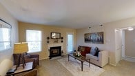 This single-floor residence offers a warm and comfortable living environment centered around a well-appointed living room. The space features a classic white-mantled fireplace that serves as the focal point of the room, flanked by decorative accessories and framed artwork above. Warm, neutral-toned walls complement the soft, plush carpeting underfoot, creating an inviting atmosphere throughout. Natural light fills the room through multiple windows dressed with white blinds, lending a bright and airy feel to the interior. The living area comfortably accommodates a sofa ensemble arranged around a rustic wooden coffee table, anchored by a large area rug. A bold, colorful canvas painting adds a vibrant accent to the neutral palette of the walls. Ceiling-mounted HVAC vents are integrated into the layout, ensuring year-round climate comfort. The layout is practical and functional, with a door providing additional access or egress from the living space. Overall, the residence combines classic architectural details with a cozy, livable design that makes it an approachable and comfortable home.