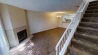 This two-story residence offers a well-organized layout designed for comfortable everyday living. The main level centers around a spacious open-concept living area finished with rich hardwood flooring that extends throughout. A tile-surround fireplace with a classic hearth serves as a natural focal point, adding warmth and character to the room. The textured ceiling adds a traditional architectural touch, while neutral wall tones create a clean, versatile backdrop. An elegant chandelier illuminates the living space and draws the eye toward the adjoining kitchen, which is visible through a pass-through opening. The kitchen features white cabinetry, dark countertops, and tiled flooring, offering a functional and bright workspace. A white-railed staircase with carpeted treads connects the main level to the upper floor, providing a clear visual division between the living and private areas of the home. The upper level offers additional living or sleeping quarters, completing the two-story floor plan. Throughout the space, the combination of hardwood floors, neutral finishes, and classic fixtures contributes to a cohesive and inviting atmosphere suited for a range of lifestyles.
