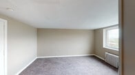 This single-floor unit offers a straightforward and functional layout spread across several rooms. The living areas and bedrooms are finished with wall-to-wall carpet in neutral grey tones, complemented by warm beige walls and clean white baseboards that create a simple, cohesive aesthetic throughout. The rooms are generously proportioned, providing ample open floor space, and several windows allow natural light to filter in, contributing to a bright and airy feel.

The kitchen is compact yet practical, featuring white cabinetry overhead, a coil-burner electric range, a single-basin sink with a wood-toned countertop, and a window that brings in natural light and exterior views. The layout is efficient, making good use of the available space.

The bathroom includes classic white tile wainscoting along the lower walls, a toilet, a wall-mounted mirror with a medicine cabinet, and a full tub and shower combination with chrome fixtures. A GFCI outlet is conveniently positioned near the sink area.

Additional rooms include a secondary carpeted bedroom or flexible space with light grey walls, and another room featuring a cast-iron radiator beneath a window — indicative of a traditional heating system. The 3D floorplan reveals a linear, well-segmented layout connecting all areas efficiently on a single level.