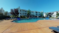 This outdoor pool area in O'Fallon, Missouri offers a well-appointed communal retreat designed for relaxation and leisure. The centerpiece is a generously sized swimming pool with clear blue water, framed by a broad, stained concrete sundeck that provides ample space for lounging and socializing. Rows of wicker-style chaise lounges with dark cushions are arranged along the deck's perimeter, complemented by additional blue lounge chairs and shaded seating beneath a striped umbrella. A decorative water feature is integrated into the pool's design, adding visual interest and ambient sound.

The pool area is surrounded by mature trees, ornamental grasses, and neatly trimmed hedgerows that enhance privacy and create a resort-like atmosphere. Multi-story residential buildings with balconies form the backdrop, reinforcing the community setting.

Adjacent to the pool is a clubhouse entrance characterized by classic architectural details, including white columns, brick accents, large arched windows, and bold red French doors. Concrete steps with black iron railings lead up to the entrance, and a white rocking chair on the porch adds a welcoming touch. The overall environment is bright and open, benefiting from abundant natural sunlight throughout the day.