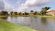 This single-story residence is set within a well-established community characterized by its tranquil, park-like environment. The most notable exterior feature is the expansive pond that serves as a central focal point of the community's landscape, reflecting the open sky and surrounded by carefully maintained green lawns. Mature palm trees and native Florida vegetation frame the buildings, contributing to a lush, tropical ambiance that is both visually appealing and naturally calming.

The residential buildings within the community display a traditional architectural style, featuring warm earth-toned exteriors with tiled rooflines that complement the natural surroundings. Exterior staircases and covered walkways add to the community's layered, connected design.

The interior images provided are insufficiently lit to allow for detailed description of the interior spaces. However, the single-floor layout suggests an accessible, straightforward floor plan designed for comfortable, everyday living. The community's outdoor setting, with its waterfront views and abundant landscaping, likely enhances the overall living experience, offering residents a peaceful natural backdrop and a strong sense of place within a verdant, Florida-style environment.