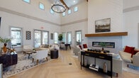 This thoughtfully designed single-floor community clubhouse blends modern farmhouse aesthetics with upscale amenities throughout its expansive layout. The centerpiece of the space is a grand main lounge with dramatically soaring ceilings accented by exposed wood beams, a striking wrought-iron chandelier, and a herringbone-patterned hardwood floor. Clerestory windows line the upper walls, flooding the room with natural light. A sleek linear fireplace with a wood-mantel surround anchors one wall, while multiple seating arrangements create inviting gathering areas. Large glass-paned doors open outward, connecting the interior to outdoor views. A secondary lounge area offers additional casual seating with expansive windows overlooking the surrounding grounds. The facility includes a dedicated conference or meeting room with a round table, leather chairs, tiled wet bar, and a beverage cooler. A work or co-working nook with a long white desk overlooks the fitness center through interior windows. The kitchenette is well-appointed with gray cabinetry, white countertops, a decorative tile backsplash, microwave, coffee station, sink, and mounted display. Practical amenities include a package locker station, dual water fountain with bottle-filling station, and a billiards room. A sliding barn door adds a refined rustic touch to the overall design.