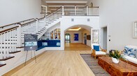 Located at 898 Water Tower Way in Lantana, Florida, this coastal-inspired leasing office makes a striking first impression with its dramatic double-height entry and grand staircase. The main lobby showcases wide-plank hardwood flooring, crisp white shiplap walls, and a sweeping staircase with dark wood treads and white balusters that rises to an open upper-level mezzanine. A built-in bench with navy blue upholstery and integrated shelving nestled beneath the staircase adds both function and charm.

A graceful arched passageway connects the entry to the main interior corridor, where tray ceilings with warm accent lighting guide visitors deeper into the space. The leasing reception area features navy blue desks set against a decorative shiplap accent wall adorned with brass sconce lighting and an elegant oval geometric pattern. Striped jute and navy area rugs complement the rattan seating with white cushions, reinforcing the sophisticated coastal aesthetic throughout.

A welcoming lounge area near the entry offers floor-to-ceiling gridded windows that flood the space with natural light, paired with rattan furniture, tropical-print throw pillows, and a striking live-edge wood coffee table. Vibrant blue abstract artwork punctuates the walls, tying the color palette together. The overall ambiance is refined yet relaxed, combining coastal Florida charm with polished, professional design elements.