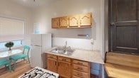This single-floor space blends classic character with functional living. The bedroom is a highlight, offering warm hardwood floors, dark-stained wood trim, and a ceiling fan with light fixture. A generously sized triple window floods the room with natural light, while a secondary window adds additional brightness. A comfortable sitting area within the bedroom provides a relaxing nook, complete with a loveseat, accent rug in vibrant teal tones, and ample space for a seating arrangement. An open closet offers convenient storage with built-in hanging rods.

The bathroom retains its vintage charm with a striking clawfoot tub fitted with a wraparound shower curtain rod, a classic pedestal sink, a dark-framed medicine cabinet mirror, and classic white hexagonal tile flooring. A window and a cast-iron radiator complete the room's timeless character.

The kitchen features warm oak cabinetry with arched detail, light stone-look countertops, a double stainless steel sink, and a gas range. A full-size refrigerator rounds out the functional layout. Throughout the space, consistent hardwood flooring and dark wood trim create a cohesive, classic aesthetic that speaks to the enduring craftsmanship of the original construction.